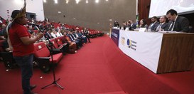 Em primeiro plano à esquerda. Um homem indígena, de perfil, fala ao microfone. Ele veste uma cam...