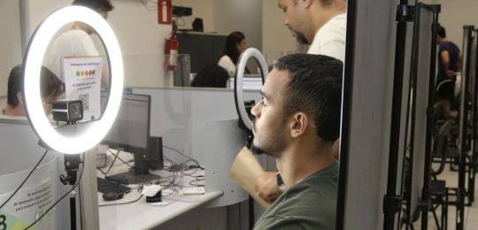 Foto mostra um homem branco com o dedo indicador sobre um leitor biométrico. A imagem da digital...