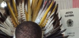 A imagem mostra, em primeiro plano, a parte de trás da cabeça de uma pessoa, que usa um cocar in...