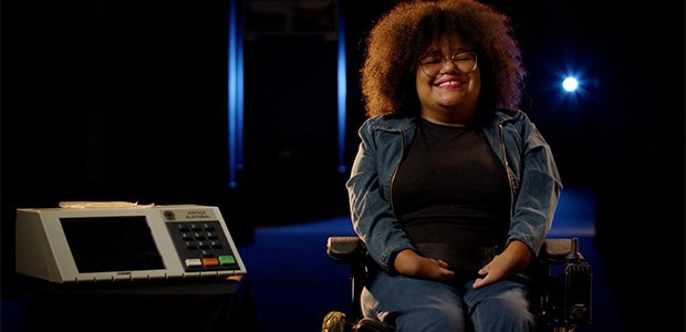 Mulher negra ,jovem e cadeirante ao lado da urna eletrônica de votação